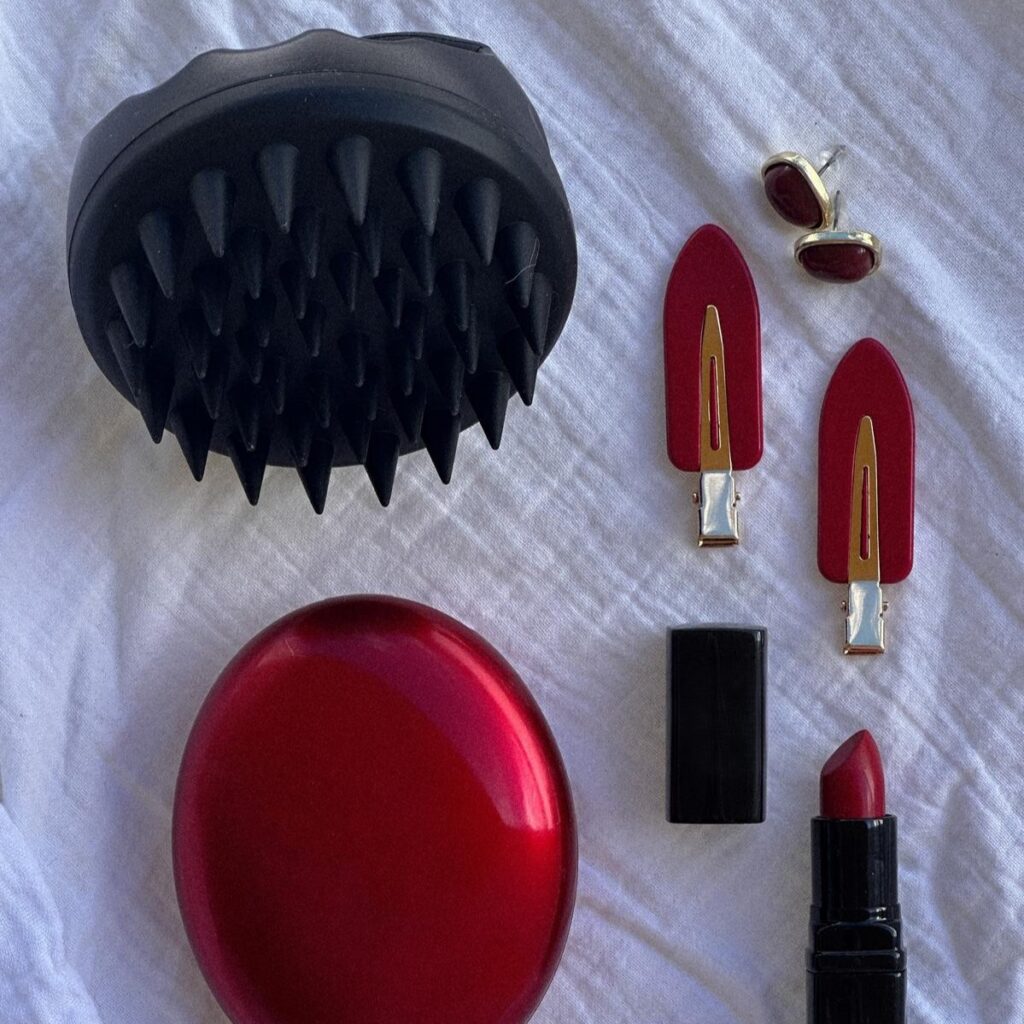 Czarna szczotka Sister Young AURA Black na jasnej tkaninie obok kosmetyków do makijażu i czerwonych akcesoriów.