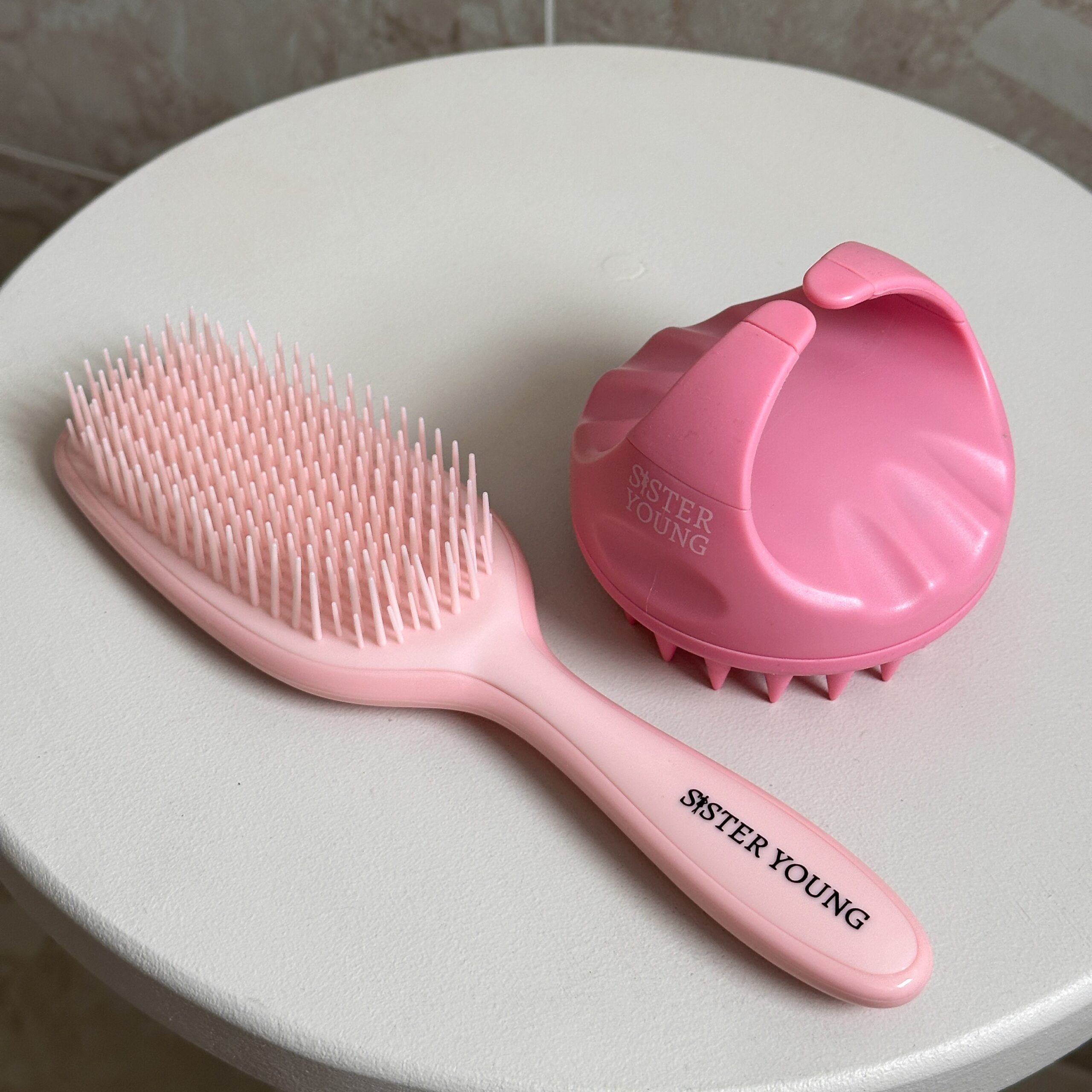 Szczotka do włosów Sister Young AYLA Pastel Peach obok różowego masażera skóry głowy AURA na jasnej powierzchni.