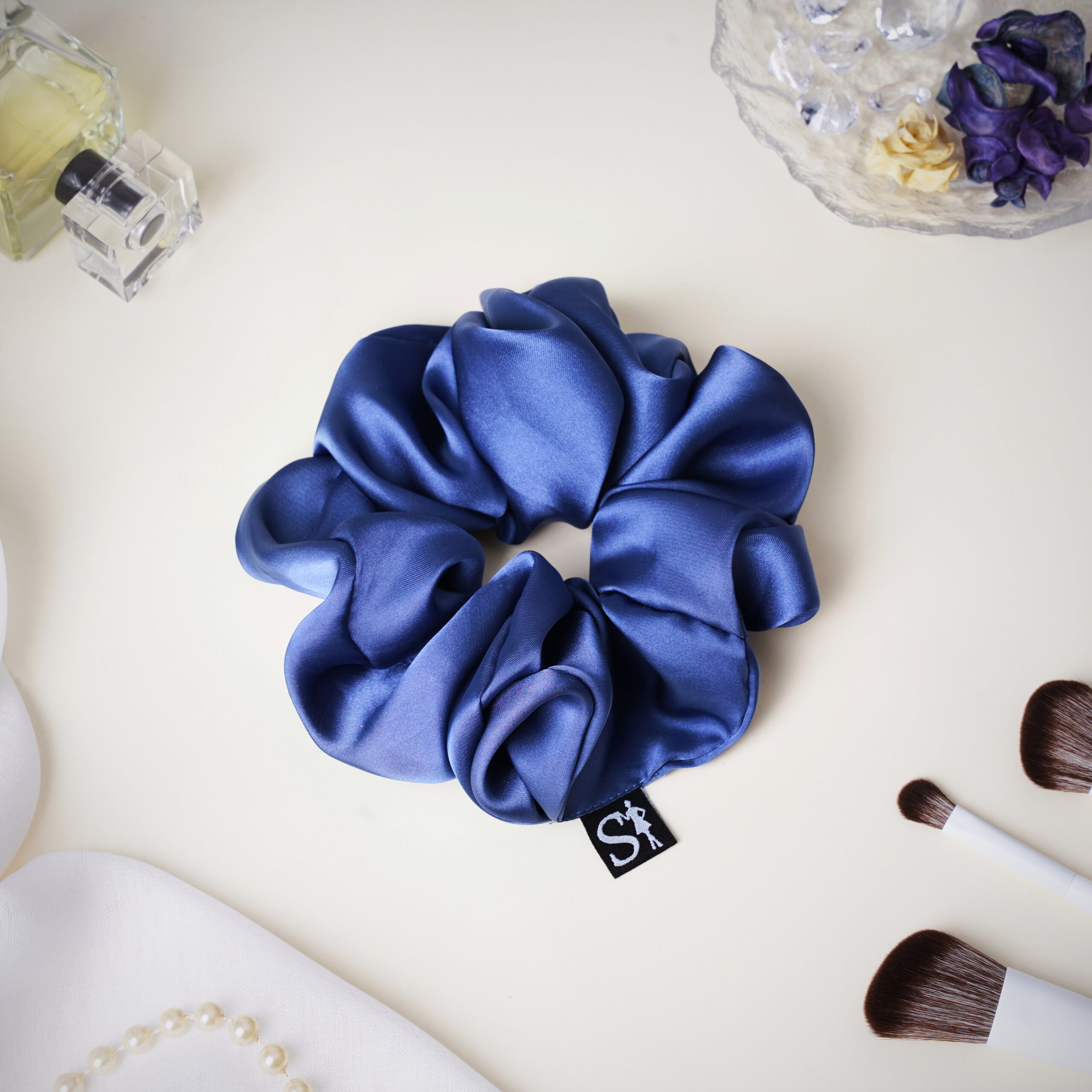 Gumka LILU Steel Blue na jasnym blacie, obok perfum, pędzli do makijażu i biżuterii.